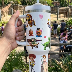 May include: White insulated tumbler featuring colorful cartoon character decals. The tumbler has a white handle, lid, and straw. The characters include a dog, Mr. Potato Head, Buzz Lightyear, and others. The tumbler is held by a person.