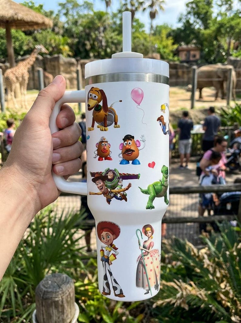 May include: White insulated tumbler featuring colorful cartoon character decals. The tumbler has a white handle, lid, and straw. The characters include a dog, Mr. Potato Head, Buzz Lightyear, and others. The tumbler is held by a person.