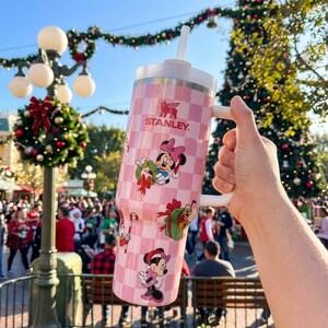 May include: Pink Stanley tumbler with a white handle and lid, featuring a checkered pattern and Minnie Mouse holiday graphics. The tumbler has a straw and the Stanley logo. The background shows a Christmas tree and festive decorations.