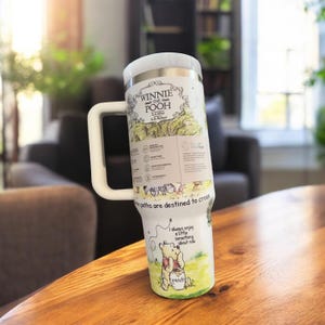 K&ouml;nnte beinhalten: Ein wei&szlig;er Reisebecher aus Edelstahl mit Illustrationen und Zitaten von Winnie Puuh. Der Becher hat einen gro&szlig;en Griff und einen wei&szlig;en Deckel. Das Design enth&auml;lt den Text "WINNIE THE POOH" und "paths are destined to cross".