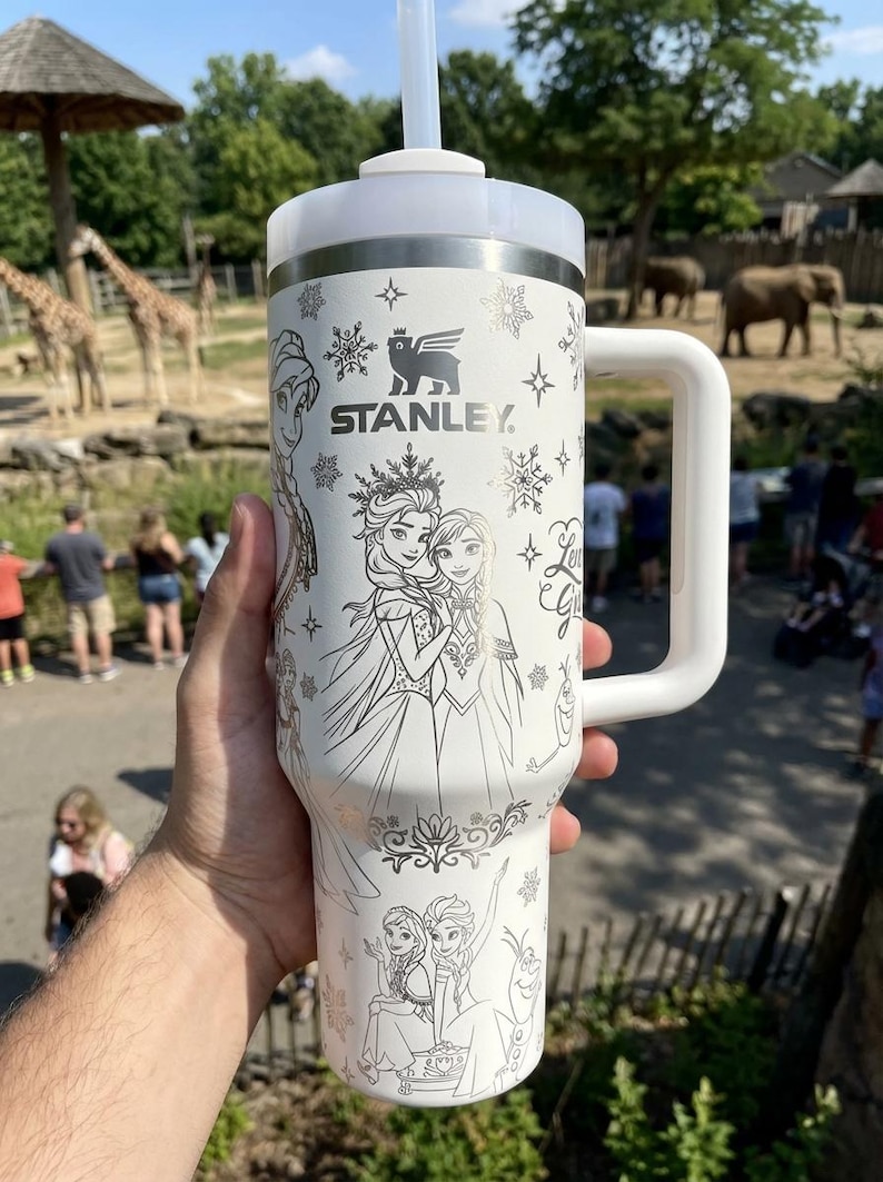 May include: White Stanley travel mug with a handle, featuring a black and white illustration of characters from the movie Frozen, snowflakes, and the Stanley logo. The mug has a white lid and a reusable straw.