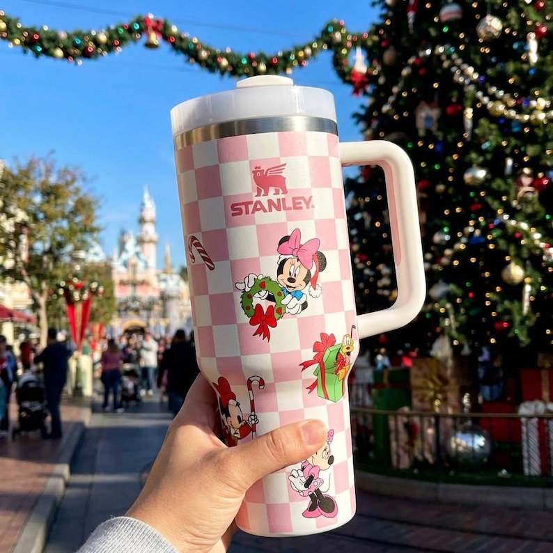 May include: A pink and white checkered Stanley tumbler featuring Minnie Mouse and holiday-themed graphics. The tumbler has a white handle and lid, with the Stanley logo visible. The design includes images of Minnie Mouse with a wreath, gifts, and candy canes.