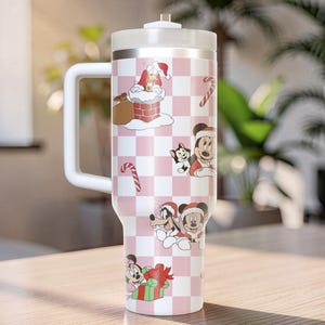 May include: A white and pink insulated tumbler with a handle, featuring a Christmas-themed design. The tumbler is decorated with cartoon characters, candy canes, and Santa Claus. The design is set against a checkered background.