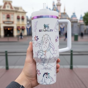 May include: White Stanley travel mug with a handle, straw, and lid, decorated with a colourful Eeyore design. The mug has the Stanley logo and floral accents. The handle is white. The background shows a blurred outdoor scene.