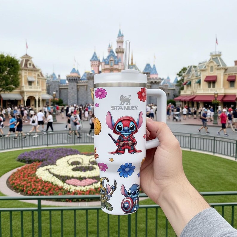 May include: White Stanley tumbler featuring a Stitch character design with a handle and straw. The tumbler has a white lid and is decorated with colorful flowers and other Stitch-themed graphics. The Stanley logo is visible on the front.