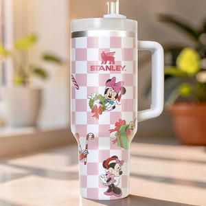 May include: A white and pink Stanley tumbler with a handle, featuring a checkered pattern and Minnie Mouse holiday-themed graphics. The tumbler has a white lid and straw, and the Stanley logo is visible.