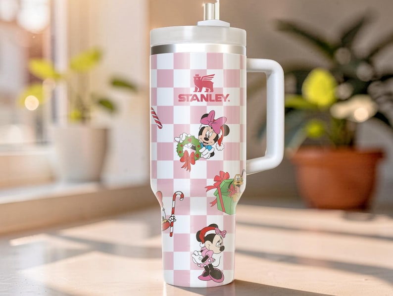 May include: A white and pink Stanley tumbler with a handle, featuring a checkered pattern and Minnie Mouse holiday-themed graphics. The tumbler has a white lid and straw, and the Stanley logo is visible.