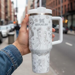 May include: White Stanley travel mug with a handle, decorated with line art of Frozen characters. The mug has a white lid and straw, the Stanley logo, and snowflake patterns. The mug is held in front of a blurred city street.