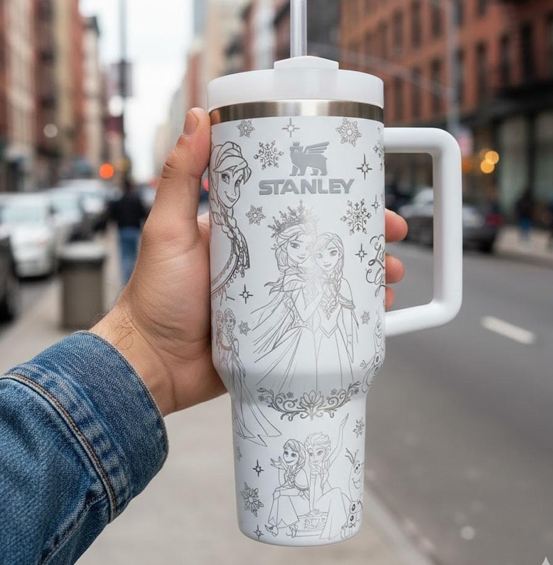 May include: White Stanley travel mug with a handle, decorated with line art of Frozen characters. The mug has a white lid and straw, the Stanley logo, and snowflake patterns. The mug is held in front of a blurred city street.