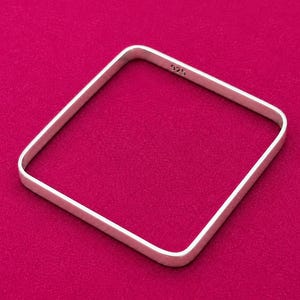 May include: A square-shaped silver bangle bracelet with rounded corners. The metal band has a smooth, matte finish and features the hallmark stamp 925 clearly visible on the inner surface, indicating the sterling silver composition of the jewelry.