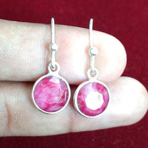 Może przedstawiać: Para srebrnych kolczyków wiszących z okrągłymi, fasetowanymi, różowymi kamieniami szlachetnymi. Kolczyki mają prosty design z małym detalem w kształcie kuli nad kamieniem szlachetnym. Tło jest ciemnoczerwone.