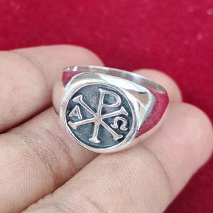 Può includere: Un anello con sigillo d'argento con un emblema circolare con il simbolo Chi Rho, un monogramma delle prime due lettere della parola greca per Cristo. L'anello ha una finitura lucida e una fascia arrotondata.