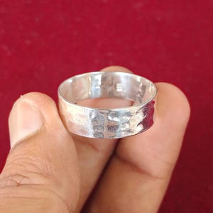 Può includere: Un anello d'argento con una texture martellata. L'anello è largo e ha un design semplice ed elegante. L'anello è tenuto in mano su uno sfondo rosso.