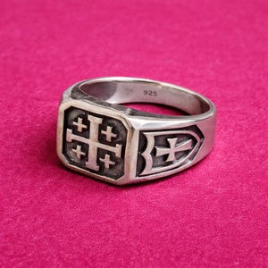 Może przedstawiać: Srebrny pierścionek z kwadratową powierzchnią z motywem krzyża jerozolimskiego. Pierścionek ma polerowane wykończenie i emblemat w kształcie tarczy z krzyżem z boku. Pierścionek jest na różowym tle.