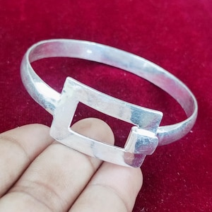 May include: A silver bangle with a rectangular clasp. The bangle has a simple, elegant design, with a smooth, polished finish. The clasp is a geometric shape, adding a modern touch. The bangle is shown against a red background.