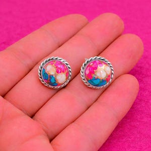 May include: A pair of round stud earrings with a silver rope-like border. The earrings feature a colorful stone with pink, turquoise, and white hues. The earrings are displayed on a pink background.
