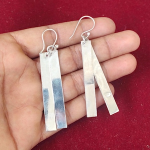 May include: A pair of silver earrings with a rectangular bar design. The earrings have a hook closure and are held in a hand against a red background. The earrings are made of a shiny, reflective metal.