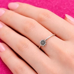 May include: A silver ring featuring a small sunflower design. The ring has a thin band and a detailed flower with textured petals and a central cluster. The ring is shown on a hand with pink nail polish against a pink background.