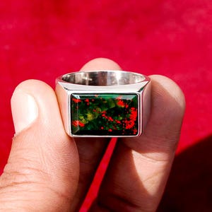 May include: A silver ring with a rectangular, green gemstone speckled with red. The ring is held against a red background, showcasing the polished metal and the stone's vibrant colors. The gemstone has a unique pattern.