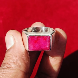 Op de afbeelding: Een zilveren ring met een rechthoekige, helder roze edelsteen. De ring heeft een dikke, gepolijste band en de edelsteen ligt gelijk met de zetting. De achtergrond is effen, dieprood.