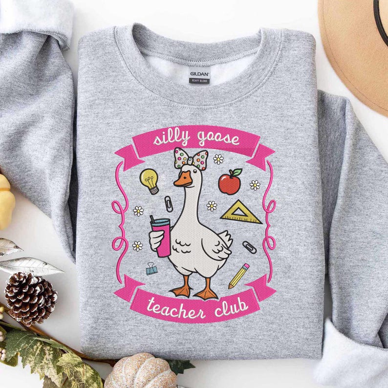 May include: A light gray sweatshirt with a whimsical graphic of a goose wearing a bow and holding a drink. The design includes the text "silly goose teacher club" in pink, along with school-themed illustrations like an apple, pencil, and ruler.