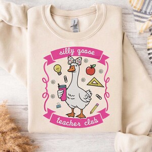 May include: Cream-colored sweatshirt featuring a cartoon goose with a bow, holding a drink, and surrounded by school-themed graphics. The text "silly goose teacher club" is displayed in pink.