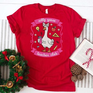 May include: A red t-shirt featuring a cartoon goose with a bow, holding a drink, and surrounded by school-themed graphics. The text on the shirt reads "silly goose teacher club".