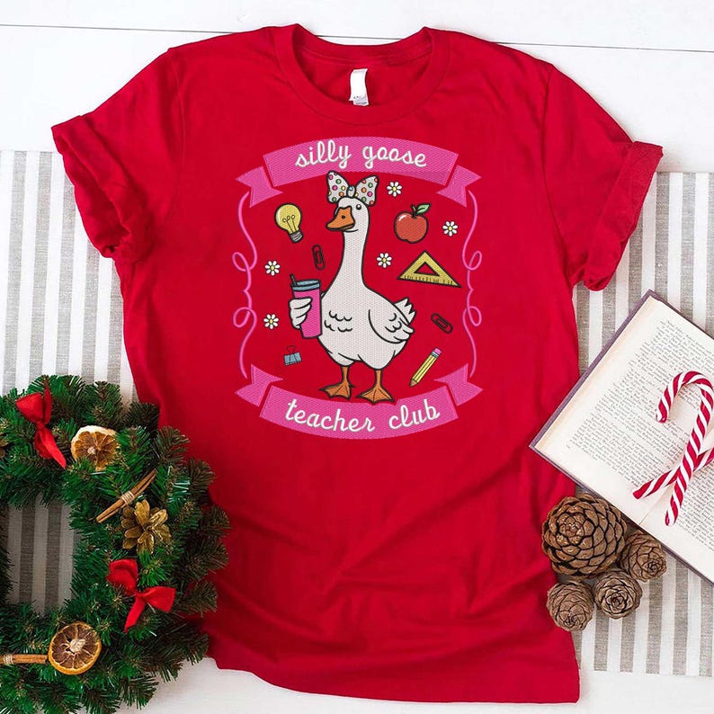 May include: A red t-shirt featuring a cartoon goose with a bow, holding a drink, and surrounded by school-themed graphics. The text on the shirt reads "silly goose teacher club".