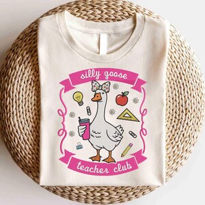 May include: Cream-colored t-shirt featuring a cartoon goose with a bow, holding a drink, and surrounded by school-themed graphics. The text "silly goose teacher club" is displayed in pink.