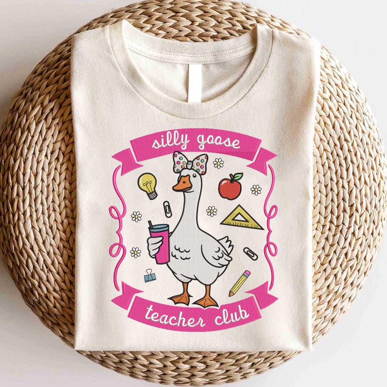 May include: Cream-colored t-shirt featuring a cartoon goose with a bow, holding a drink, and surrounded by school-themed graphics. The text "silly goose teacher club" is displayed in pink.