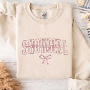 May include: A beige sweatshirt with the text "SHOWGIRL in my era" in pink lettering. A pink bow is below the text. The sweatshirt is folded on a light wood surface.