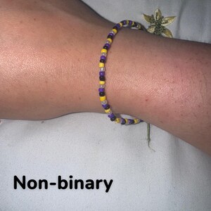May include: A bracelet made of small yellow, purple, black, and white beads. The bracelet is worn on a wrist. The image includes the text "Non-binary".