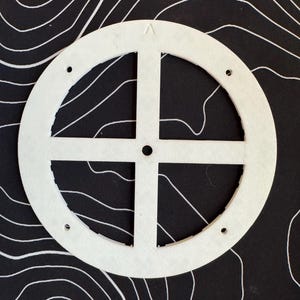 Może przedstawiać: Biały, okrągły przedmiot z wycięciem w kształcie krzyża pośrodku i trzema małymi otworami. Umieszczony na czarnym tle z białymi, zakrzywionymi liniami, tworzącymi wzór mapy topograficznej. Projekt przedmiotu jest symetryczny.