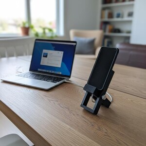 May include: A black smartphone is propped up on a black phone stand on a wooden table. A silver laptop is connected to the phone stand with a white cable. The laptop screen displays a blue and white interface.
