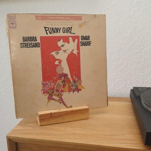 May include: A vintage vinyl record album cover for "Funny Girl" featuring Barbra Streisand and Omar Sharif. The cover has a red and white illustration of two people kissing. The album is propped up on a small wooden stand.