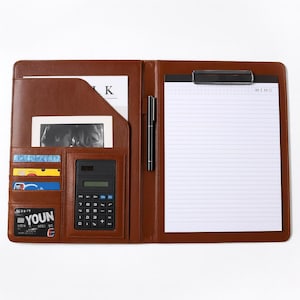 Könnte beinhalten: Offenes braunes Leder-Portfolio mit Notizblock, Stift, Taschenrechner und Kartenfächern. Das Portfolio bietet mehrere Fächer für Dokumente und Zubehör und ist für die Organisation konzipiert.