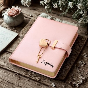 May include: A pink journal with a heart-shaped lock and key charm. The journal has a gold-colored clasp and the name "Sophia" printed on the cover. The journal is closed and sits on a wooden surface.