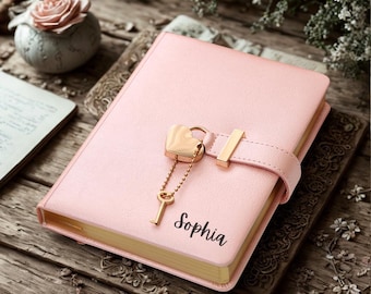 May include: A light pink journal with a gold-toned heart-shaped padlock and a matching key attached by a delicate chain. The cover features the name Sophia printed in black script. The journal has gold-edged pages and a secure strap closure.
