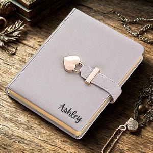 May include: A gray journal with a heart-shaped clasp and the name "Ashley" printed on the cover. The journal has a gold-colored edge and a strap closure. The journal is on a wooden surface.