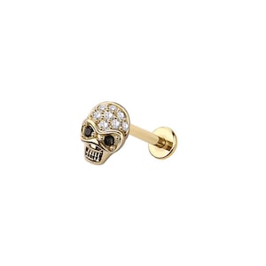 May include: A gold-colored skull-shaped earring with a flat back for piercing. The skull's eyes are black, and the forehead is covered in small, clear gemstones. The earring post is also gold.