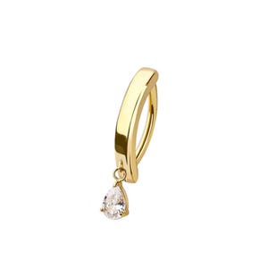 May include: A gold-colored belly button ring with a curved bar and a dangling pear-shaped clear gemstone. The jewelry is set against a white background, highlighting the ring's design and sparkle.