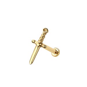 Pode incluir: Uma joia corporal dourada em forma de espada. A joia apresenta uma guarda detalhada e uma lâmina pontiaguda. A peça foi projetada para ser inserida em um piercing. O design geral é uma representação em miniatura de uma espada.