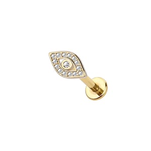 Puede incluir: Un pendiente dorado en forma de ojo con la parte posterior plana. El diseño del ojo está delineado con pequeñas piedras preciosas transparentes, y una piedra preciosa más grande está en el centro. El pendiente está diseñado para un piercing en el labio o en la oreja.