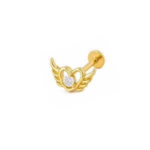 May include: A gold-toned flat back stud earring featuring a heart-shaped frame with stylized wings extending from the sides. A single clear round gemstone is set in the center of the heart. The post is visible extending from the back of the piece.