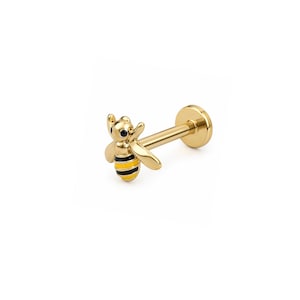 May include: A gold-tone flat back stud earring featuring a small bee charm. The bee has polished gold wings and a body decorated with horizontal black and yellow enamel stripes. The post is straight and ends in a circular flat disc backing.