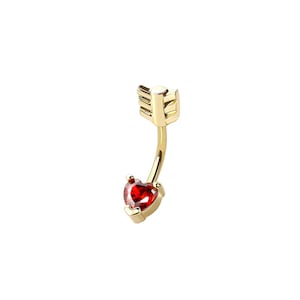 May include: A gold-colored belly button ring with a curved barbell design. The ring features a red heart-shaped gemstone at the bottom and an arrow-shaped top. The jewelry is set against a white background.