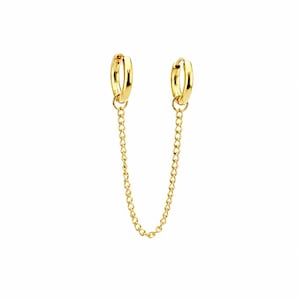 May include: A pair of gold-colored hoop earrings connected by a delicate chain. The earrings have a simple, elegant design, with a polished finish. The chain dangles between the two hoops, creating a stylish and modern look. The jewelry is set against a white background.