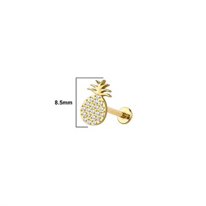 May include: A gold-colored, pineapple-shaped earring with a flat back. The pineapple is encrusted with small, clear gemstones. The earring measures 8.5mm in height. The jewelry is designed for body piercings.