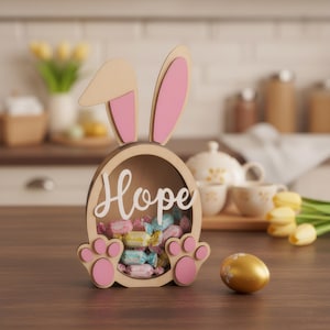 May include: Wooden Easter bunny-shaped candy holder with pink accents and the word "Hope". The bunny has pink paws and ears. The holder is filled with wrapped candies, and a gold egg is next to it.
