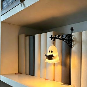May include: A white, glowing ghost-shaped lamp holding a small black book, suspended from a black, ornate, scroll-work metal wall-mount bracket. The lamp emits a warm, soft light and is positioned between several books on a white shelf.